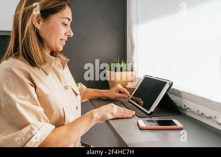Side view of mature female sitting at counter in kitchen and browsing tablet in morning Stock Photo