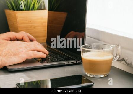 Cropped unrecognizable middle aged male working on counter with netbook and cup of coffee in kitchen in the morning Stock Photo