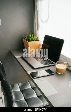 Modern laptop and smartphone placed on counter with cup of morning coffee in kitchen lit by sunlight Stock Photo