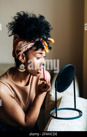 Side view of charming African American female looking in round mirror and applying powder on face while doing makeup at home Stock Photo