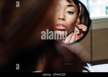 Reflection of content African American female putting on earring and looking in round mirror at home Stock Photo