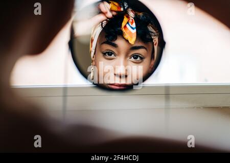 Reflection of content African American female putting on headband and looking in round mirror at home Stock Photo