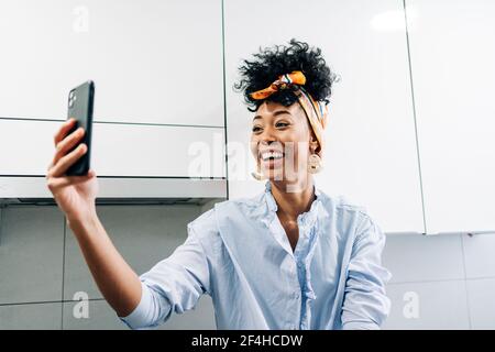 Cheerful black housewife in headband taking self portrait on smartphone in kitchen at home Stock Photo