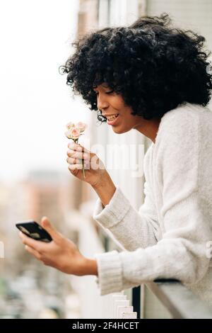 Side view of African American female standing on balcony and taking self portrait with rose flowers while using smartphone Stock Photo