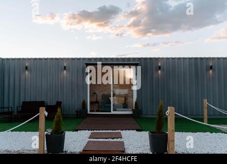 Modern gray shipping container house with glass doors located on grassy meadow and decorated with lush potted plants Stock Photo