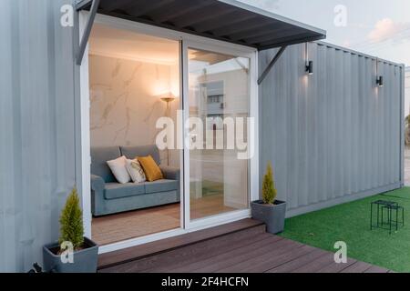 Contemporary gray container house with comfy sofa behind glass door located on grassy yard Stock Photo