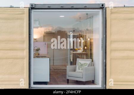 Small stylish kitchen and comfy armchair behind glass wall of contemporary container house in daylight Stock Photo