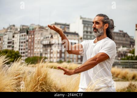 Mature unshaven ethnic male in white outfit looking forward while practicing yoga against city buildings Stock Photo