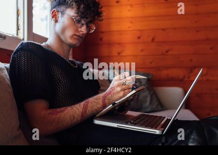Side view of crop young transsexual man taking notes in notebook against portable computer while sitting on sofa in chalet Stock Photo