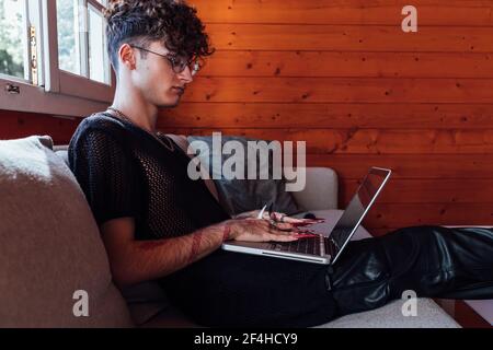 Side view of young transgender male with long nails browsing internet on netbook while resting on couch in bungalow Stock Photo