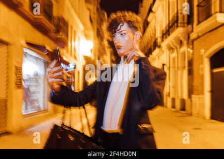 Young trendy transgender man with makeup touching cheek while taking self portrait on cellphone in town at night Stock Photo