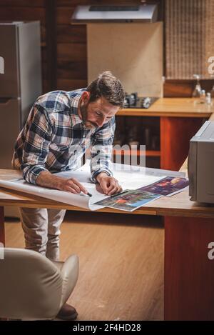 Delighted male architect standing at table and drawing plan of house on paper while working on project at home Stock Photo