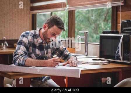 Serious male architect sitting at table in kitchen and drawing blueprint of building with pencil and ruler while working at home Stock Photo
