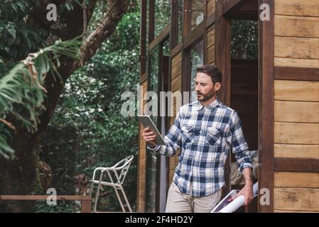 Side view of busy male architect standing on terrace with blueprint and browsing tablet while working on project Stock Photo
