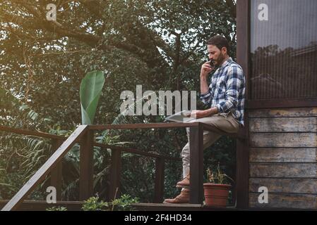 Side view of serious male architect sitting on railing on wooden terrace and talking on smartphone while discussing project Stock Photo