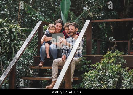 Cheerful couple with cute children sitting on wooden steps of house and taking selfie on tablet Stock Photo