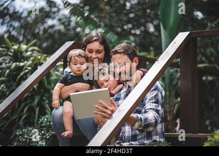 Cheerful couple with cute children sitting on wooden steps of house and taking selfie on tablet Stock Photo