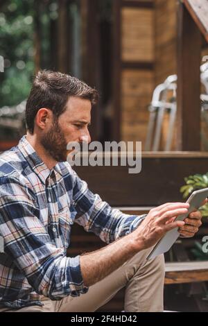 Focused male architect using tablet while sitting on wooden steps of house and working on remote project Stock Photo