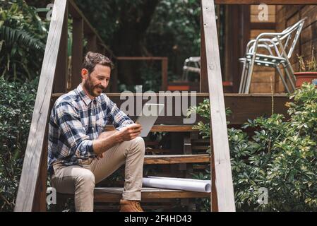 Focused male architect using tablet while sitting on wooden steps of house and working on remote project Stock Photo