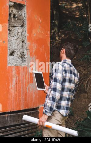 Back view of unrecognizable male architect checking on broken wall of building on tablet while working on project Stock Photo