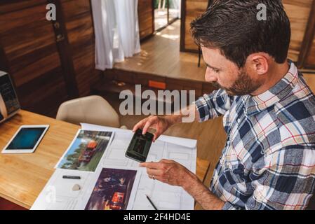 High angle side view of male architect standing at table and taking picture of draft of building while working remotely from home Stock Photo