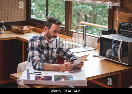Smiling male architect with blueprint of house sitting at table at home and having video call via tablet while talking about new project Stock Photo