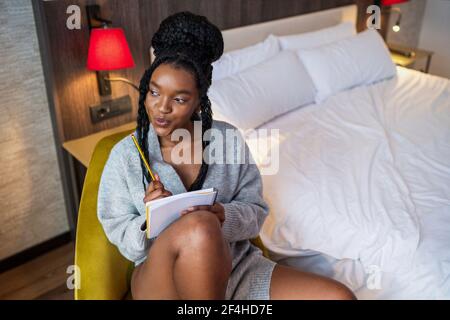 Dreamy young positive African American female millennial in casual clothes and socks sitting on comfortable armchair and looking away thoughtfully whi Stock Photo