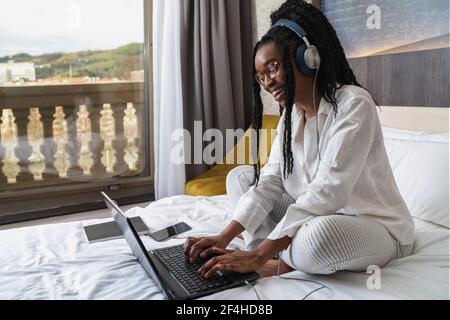 Side view of positive young African American female student in casual clothes and headphones smiling while sitting on cozy be and typing on laptop dur Stock Photo