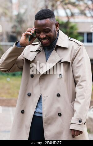 Happy adult African American male in stylish coat talking with mobile phone in hands looking away while standing on urban street in spring day Stock Photo