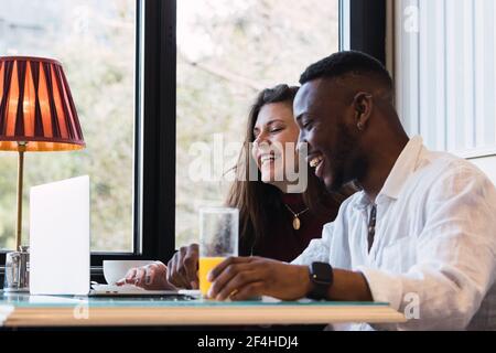 Happy multiracial couple sitting at table and browsing laptop together while having breakfast in stylish room at home Stock Photo