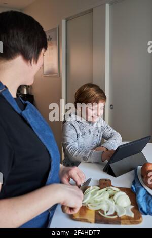 Happy boy smiling and browsing tablet near Woman cutting onion during food preparation in kitchen at home Stock Photo