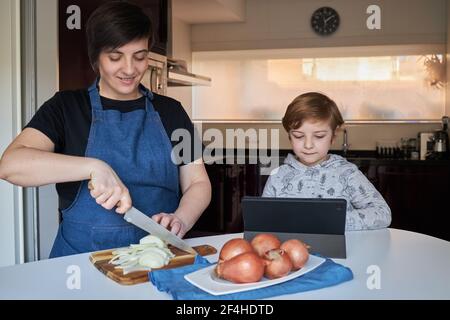 boy smiling and browsing tablet near Woman cutting onion during food preparation in kitchen at home Stock Photo