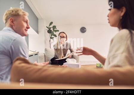 Multiethnic couple sitting on couch and talking about mental problems during therapy session with psychologist Stock Photo