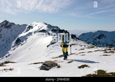 Back view of unrecognizable climber walking on slope of snow covered rocky mountain range in sunny weather Stock Photo