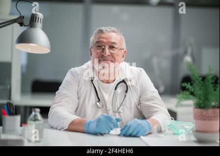 Portrait of a gray-haired doctor, who sits at a desk in the office in a dressing gown and with a stethoscope Stock Photo