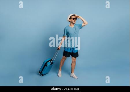 Full length caucasian joyful guy in a hat and sunglasses, dressed in summer wear, holds a blue suitcase, looks into the distance, happy about of a long-awaited vacation, isolated blue background Stock Photo