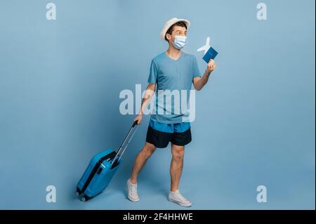 Full length caucasian joyful guy in a hat and mask, holds a blue suitcase and a small airplane with passport, looks into the distance, happy about of a long-awaited vacation, isolated blue background Stock Photo