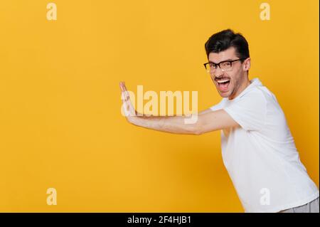 Screaming amazed excited caucasian guy wearing glasses and white basic t-shirt gesturing hands towards empty space looks at camera while standing against isolated orange background Stock Photo