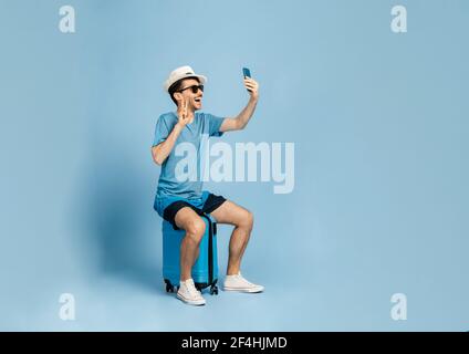 Cheerful guy dressed in summer clothes, wearing sunglasses and a hat, sits on a blue suitcase, takes a selfie on the phone or communicates on a video call, goes on vacation, blue isolated background Stock Photo