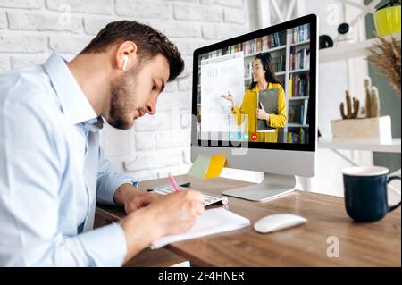Online education, e-learning. Young man, studies remotely via video call, taking notes, on the computer screen the teacher conducts an online lesson shows information on a flipchart,studying from home Stock Photo
