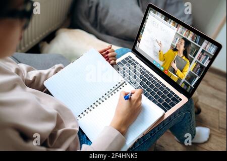 Online lecture during distance learning, on the screen female teacher explains the topic of the lesson. Girl uses a laptop, listens to an online lecture, makes notes in a notebook, studies from home Stock Photo