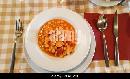 overhead view of white plate of tripe with chickpeas with cutlery with cutlery on checkered tablecloth and red napkin Stock Photo