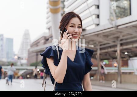 Portrait of beautiful and smiling young Asian woman in casual clothing talking to friend over smartphone while walking in city street Stock Photo