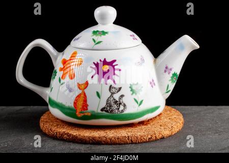 Close up isolated image of a decorative porcelain tea pot on a cork trivet against dark background. The hot pad prevents damage from hot objects onto Stock Photo
