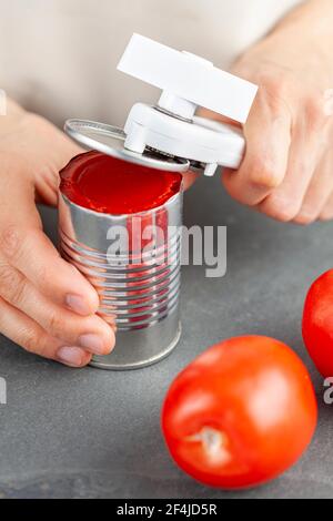 A woman is carefully opening a can of tomato paste on a kitchen counter using a white plastic can opener. She is preparing a meal for which she uses b Stock Photo