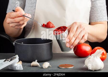 A caucasian woman is adding tomato paste from a can into a dish she is preparing in a nonstick pot.  There are fresh tomatoes, garlic, a can opener on Stock Photo