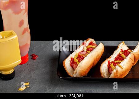 Two hot dog sandwiches made in special buns and topped with ketchup, mustard sauces are placed on a flat black plate on kitchen counter Quick and easy Stock Photo
