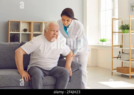 Friendly female doctor helps an older man who is trying to get up from the couch. Stock Photo