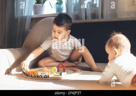 Small caucasian boy is playing on the couch with a toy train railway while newborn sibling is watching him Stock Photo