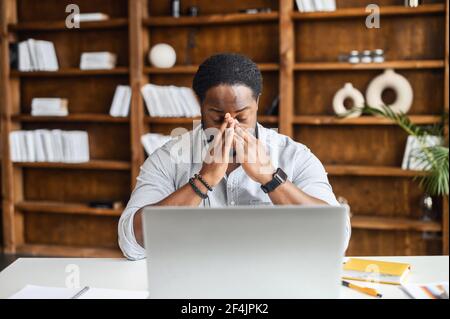 Desperate and tired young African-American businessman entrepreneur in smart casual attire, touching face, feeling stressed and overwhelmed while sitting in the modern office with open laptop Stock Photo